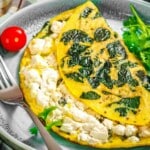 A spinach and feta omelette is folded on a plate with a cherry tomato, fresh cilantro, and a fork beside it.