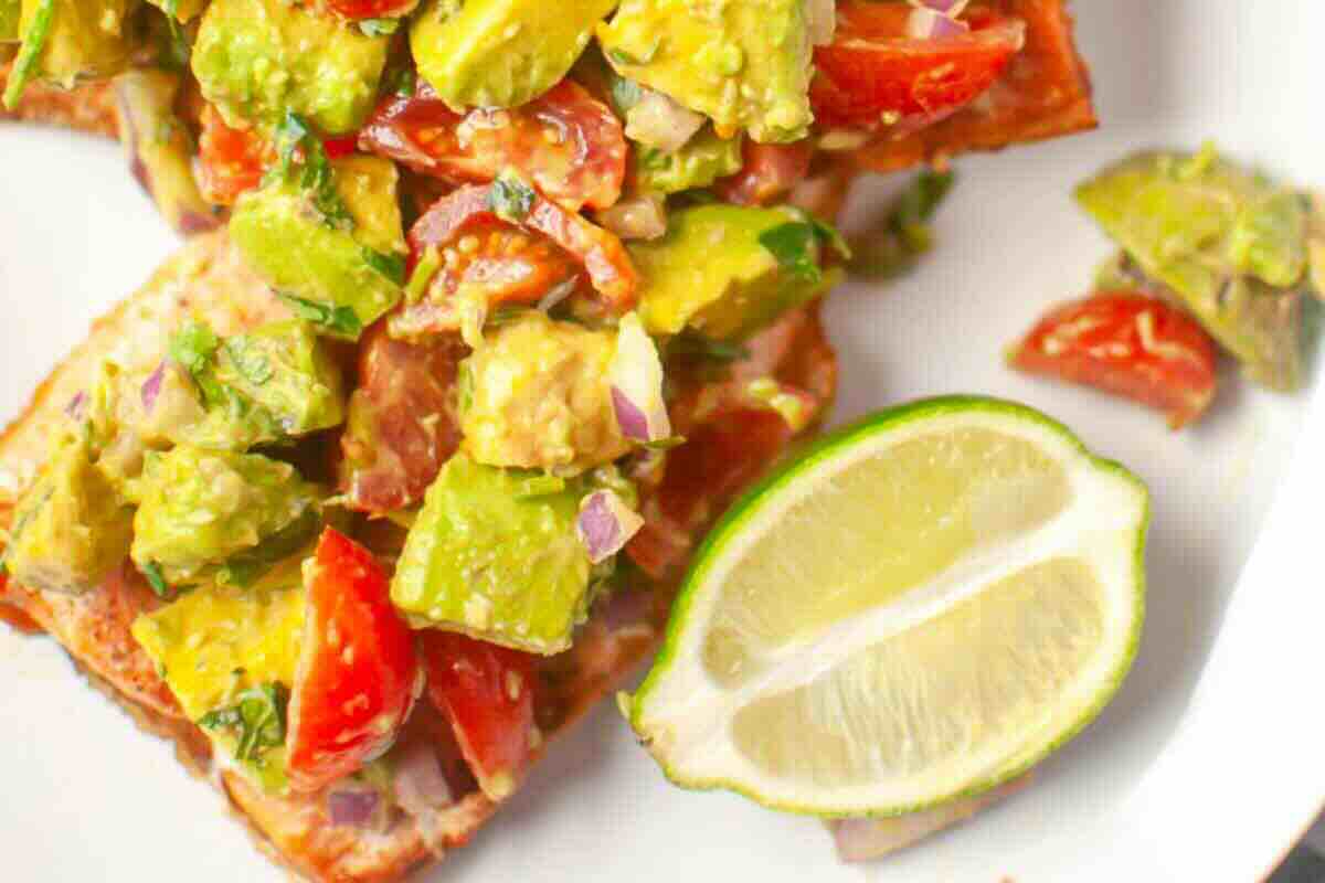 Close-up of pan-seared salmon topped with diced avocado, tomato, onion, and herbs, served with a wedge of lime on a white plate.