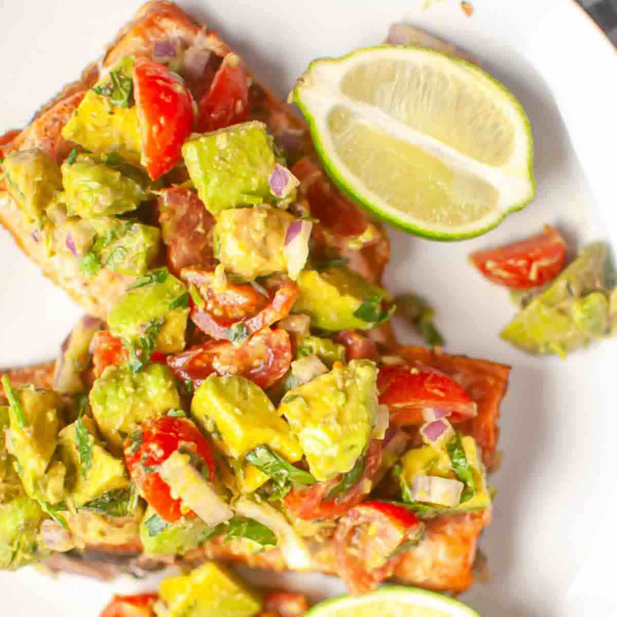 Two pieces of toast topped with a chunky avocado and tomato mixture, served with two lime wedges on a white plate.