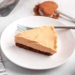 A slice of peanut butter cheesecake on a white plate with a fork, with cocoa powder and a measuring spoon in the background.
