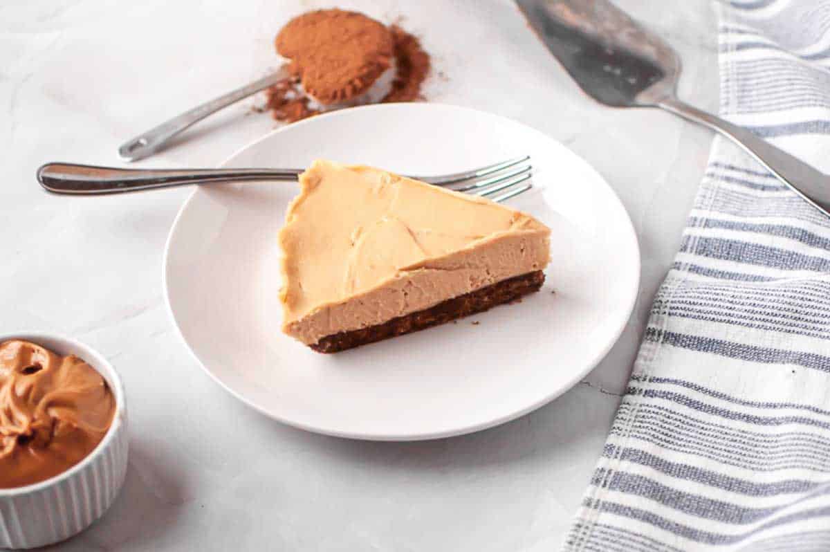 A slice of peanut butter cheesecake with a fork on a white plate, next to a bowl of peanut butter and a spoon with cocoa powder on a marble surface.