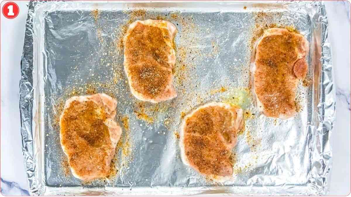 Four raw, seasoned pork chops are placed on a foil-lined baking sheet, spaced apart and ready for cooking.