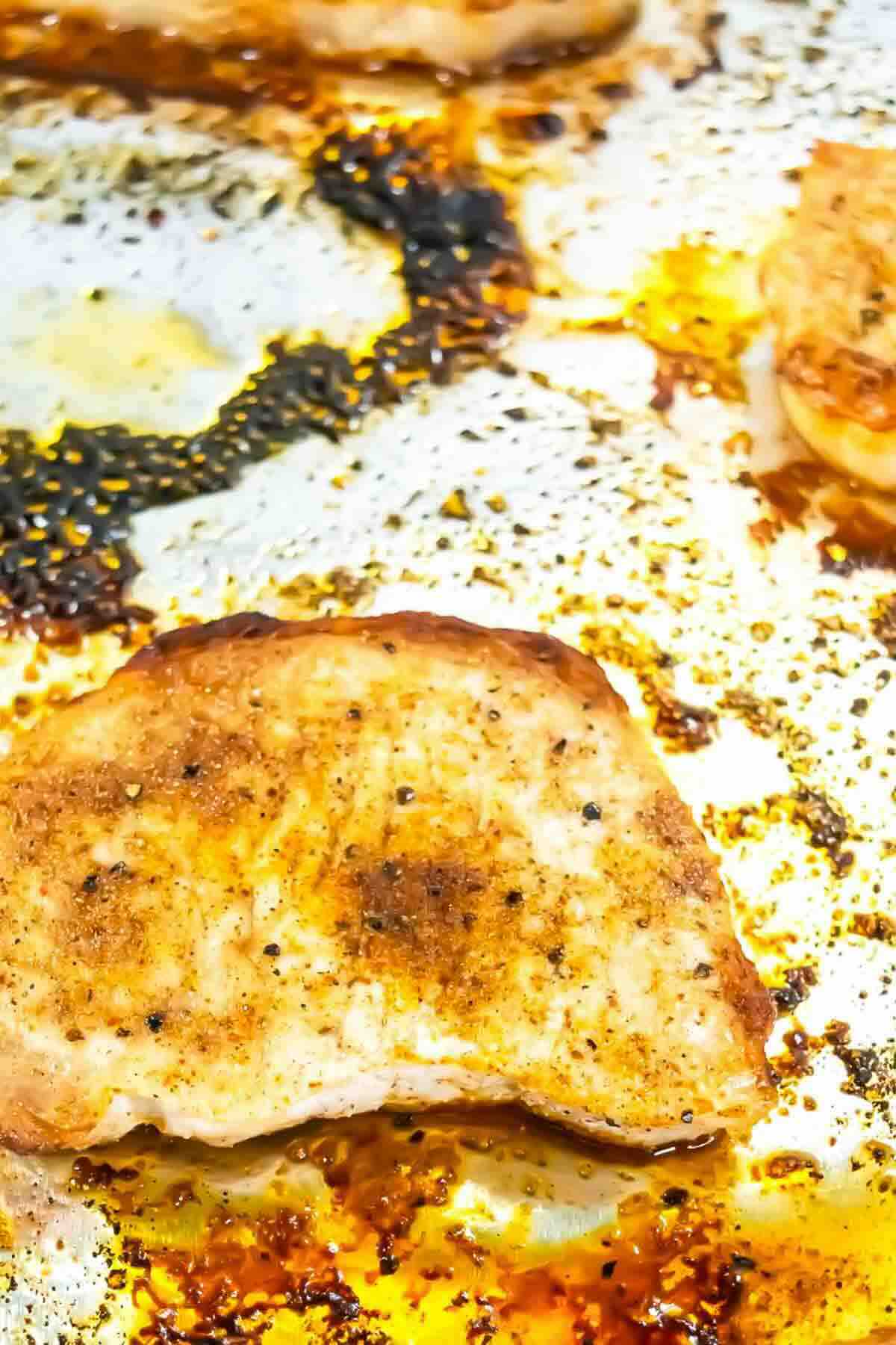 Seasoned cooked chicken breast on a baking sheet with visible oil, spices, and browned spots on the pan.