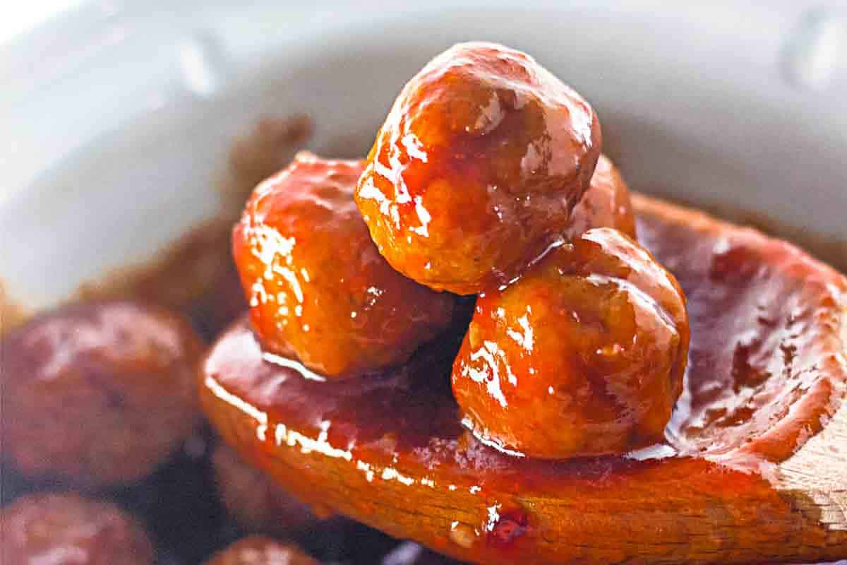 A wooden spoon holds three glazed meatballs coated in a shiny red sauce, with more meatballs and sauce visible in the background.