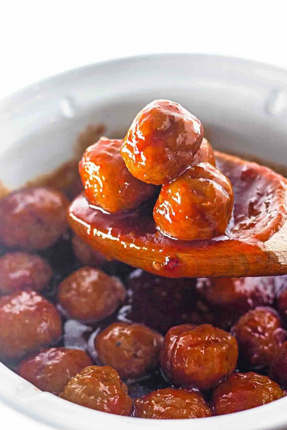 A wooden spoon lifts glazed meatballs from a slow cooker filled with sauce and more meatballs.