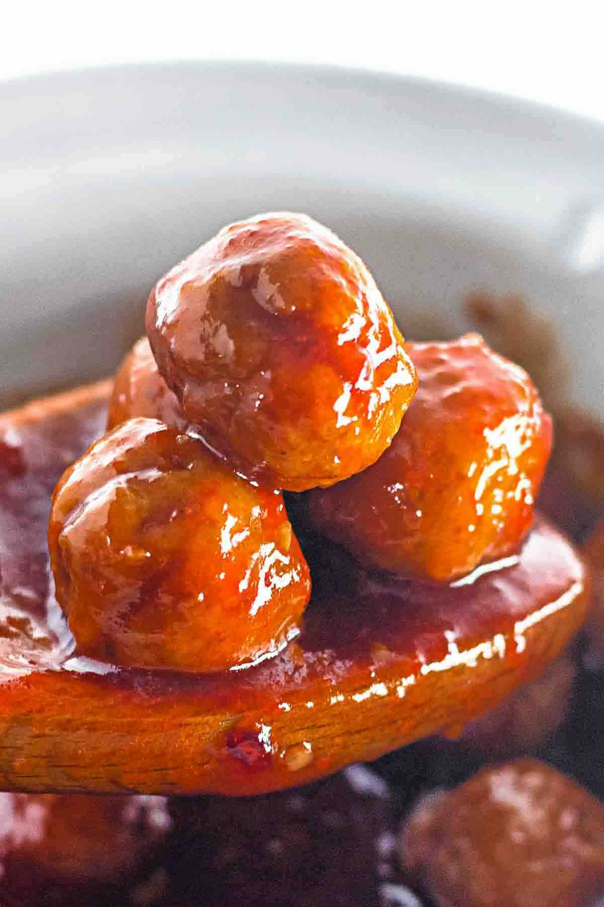 Three glazed meatballs on a wooden spoon are covered in a rich, shiny sauce, with more meatballs visible in the background.