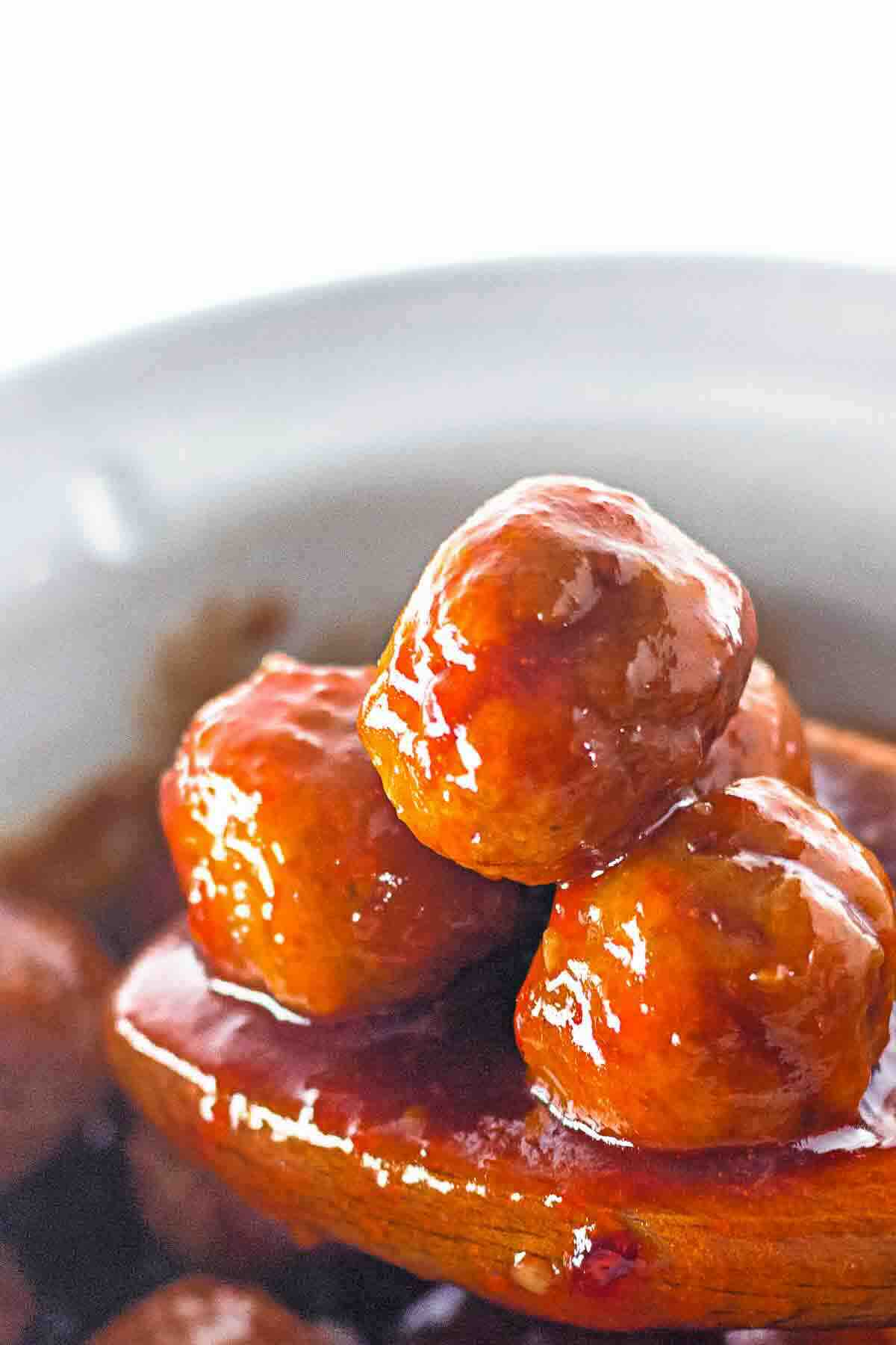 Three glazed meatballs sit on a wooden spoon above a bowl filled with more meatballs and sauce, set against a white background.