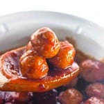 A wooden spoon holds three glossy, sauce-covered meatballs above a bowl or pot of additional meatballs.