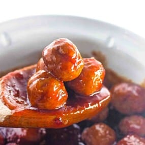 A wooden spoon holds three glossy, sauce-covered meatballs above a bowl or pot of additional meatballs.