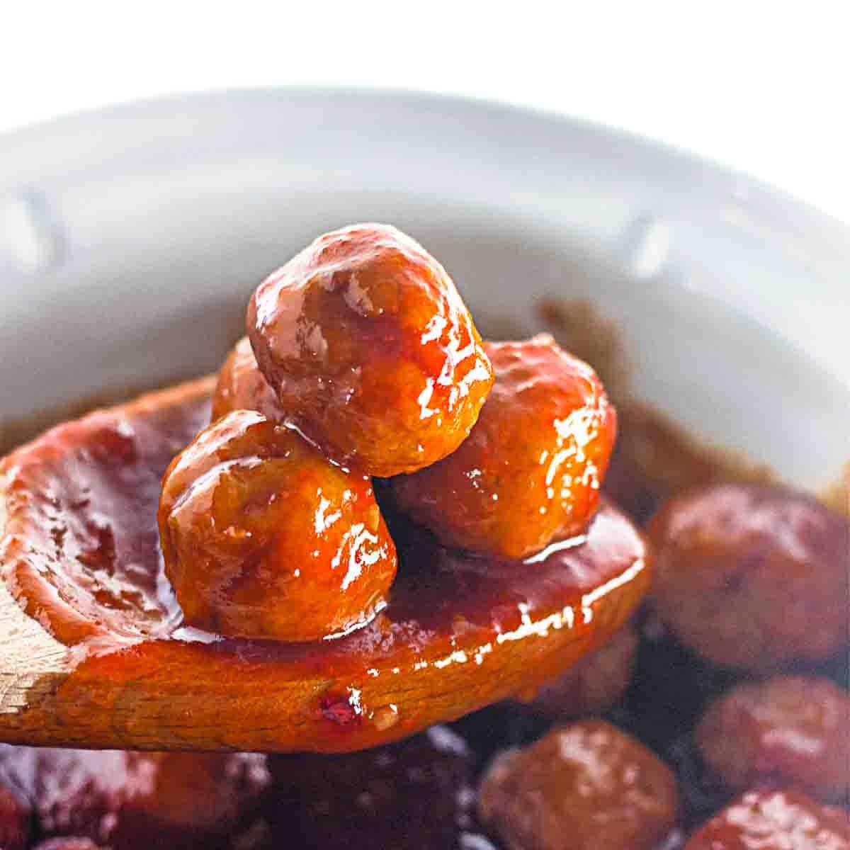 A wooden spoon holds three glossy, sauce-covered meatballs above a bowl or pot of additional meatballs.