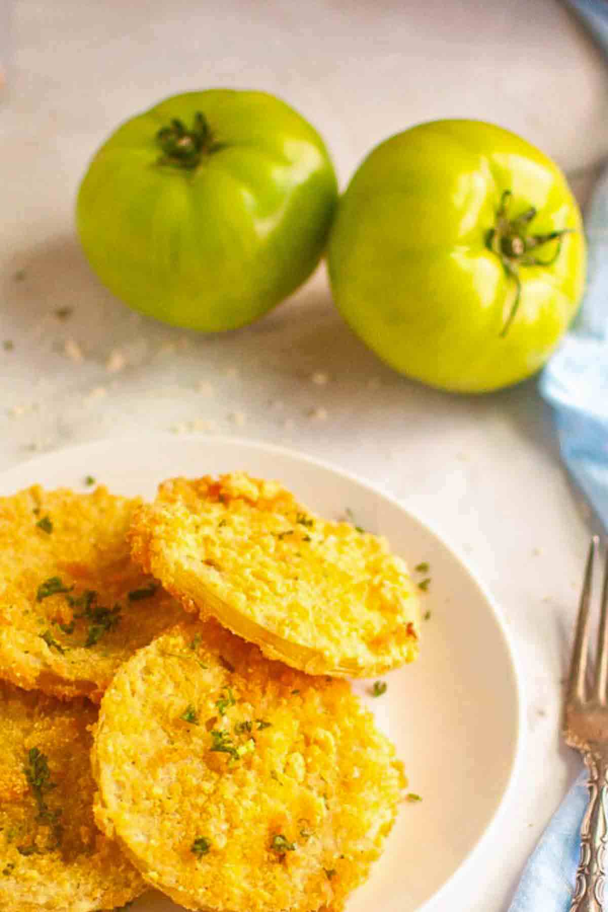 A plate of fried green tomato slices sits in the foreground, with two whole green tomatoes placed on the surface in the background.