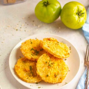 A plate of crispy fried green tomato slices garnished with herbs sits on a white surface, with two whole green tomatoes in the background.