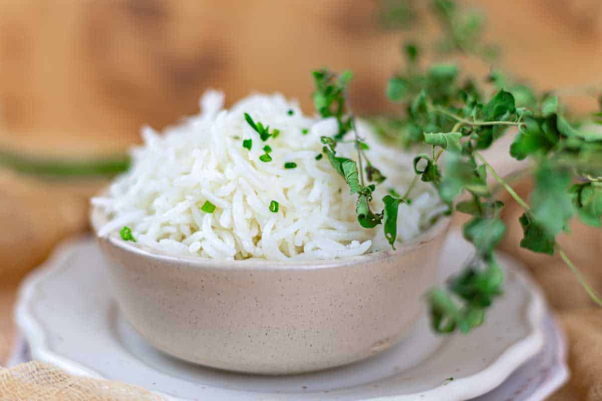 A bowl of cooked white rice garnished with chopped herbs, placed on a stack of white plates, with fresh herbs in the foreground.