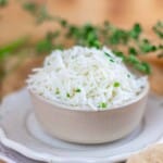 A bowl of cooked white basmati rice garnished with chopped herbs, placed on a white plate with a blurred background.