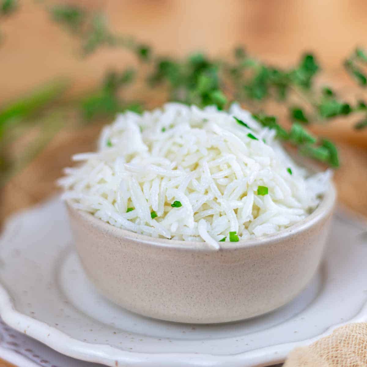 A bowl of cooked white basmati rice garnished with chopped herbs, placed on a white plate with a blurred background.