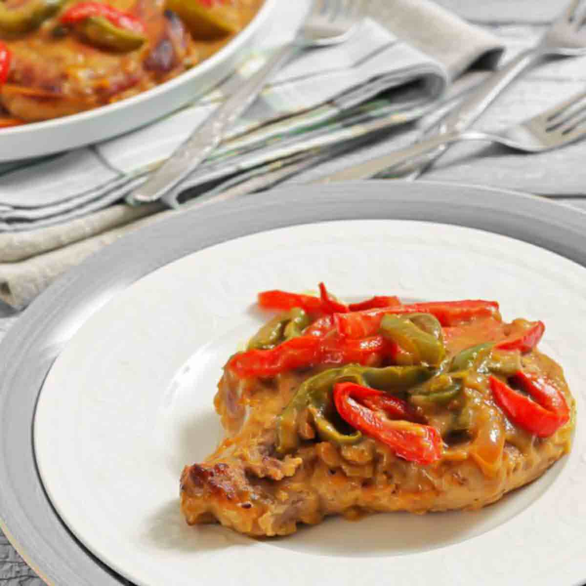 A cooked pork chop topped with sliced red and green bell peppers on a white plate, with a fork and knife in the background.
