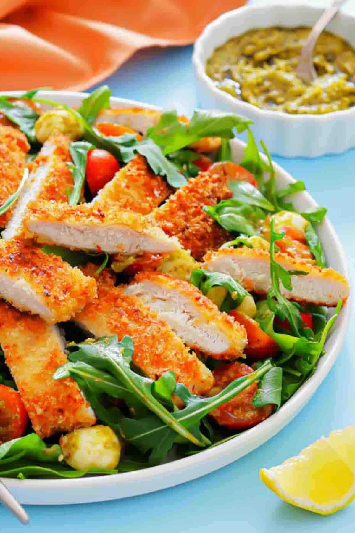 A plate of sliced breaded chicken on a bed of arugula, cherry tomatoes, and mozzarella, with a small bowl of green sauce and a lemon wedge on the side.