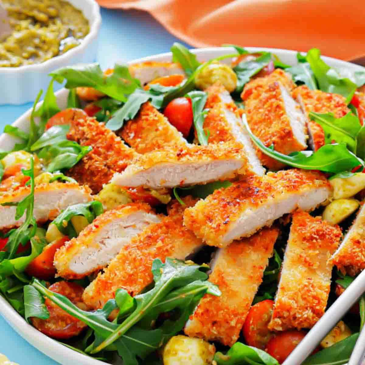 A salad with arugula, cherry tomatoes, mozzarella balls, and sliced breaded chicken, served with a side of green sauce.
