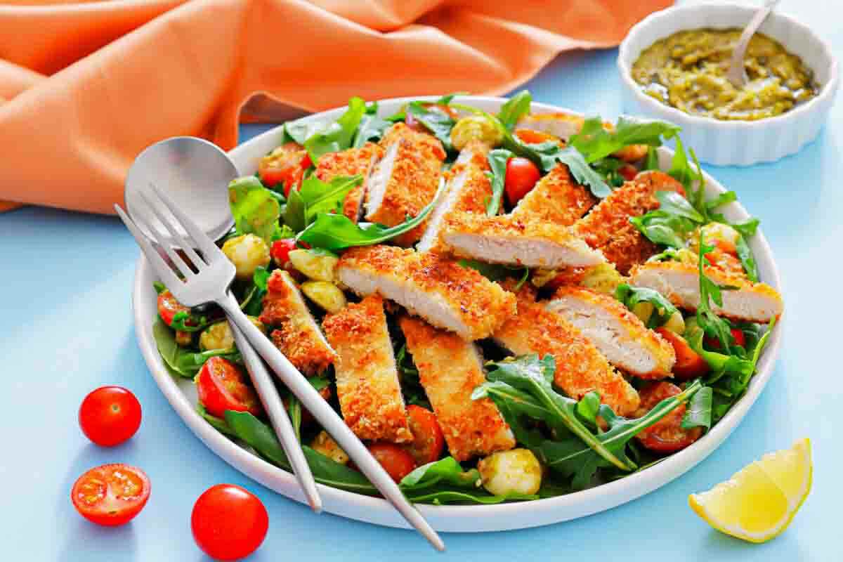 A plate of sliced breaded chicken on a bed of arugula, cherry tomatoes, and cucumbers, with a lemon wedge and a small bowl of dressing on the side.