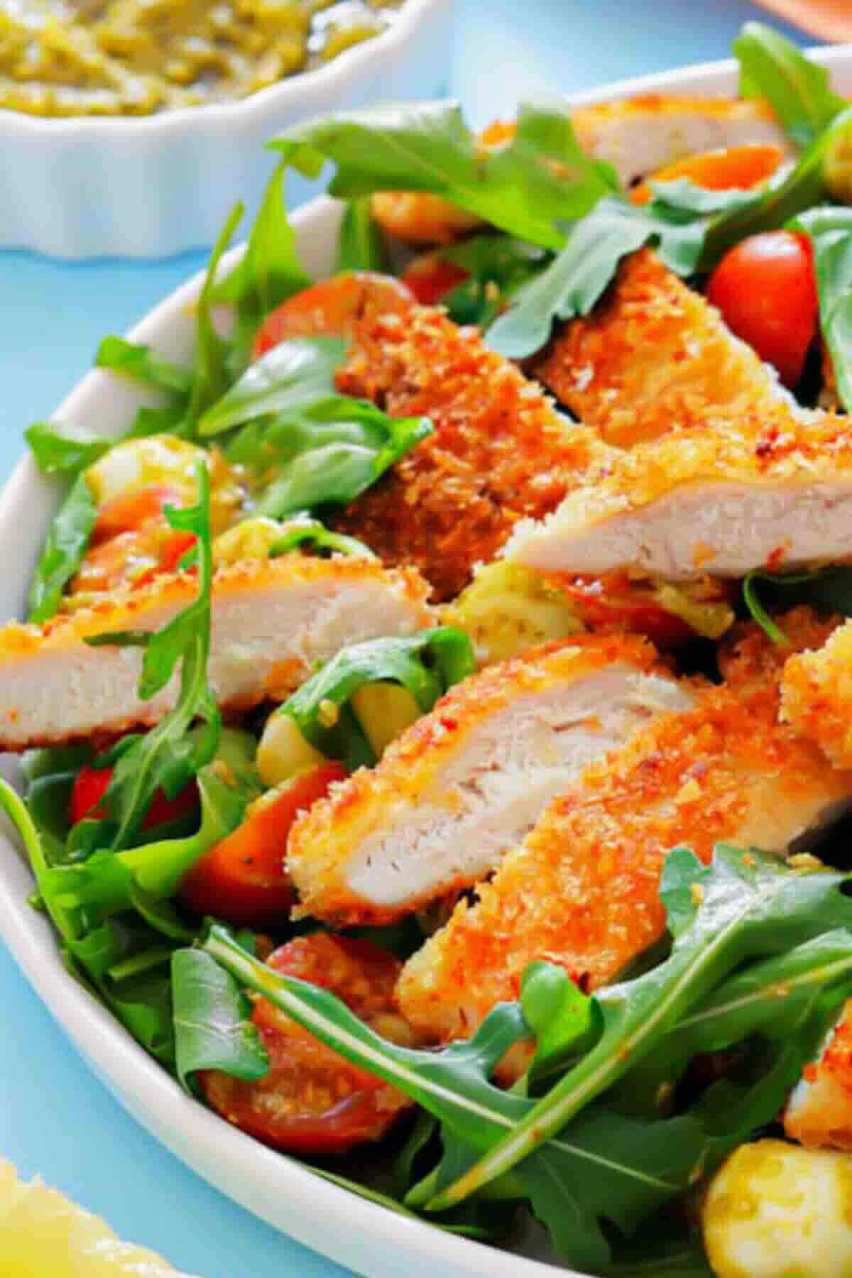 A close-up of a salad with crispy breaded chicken strips, fresh arugula, cherry tomatoes, and sliced vegetables on a white plate.