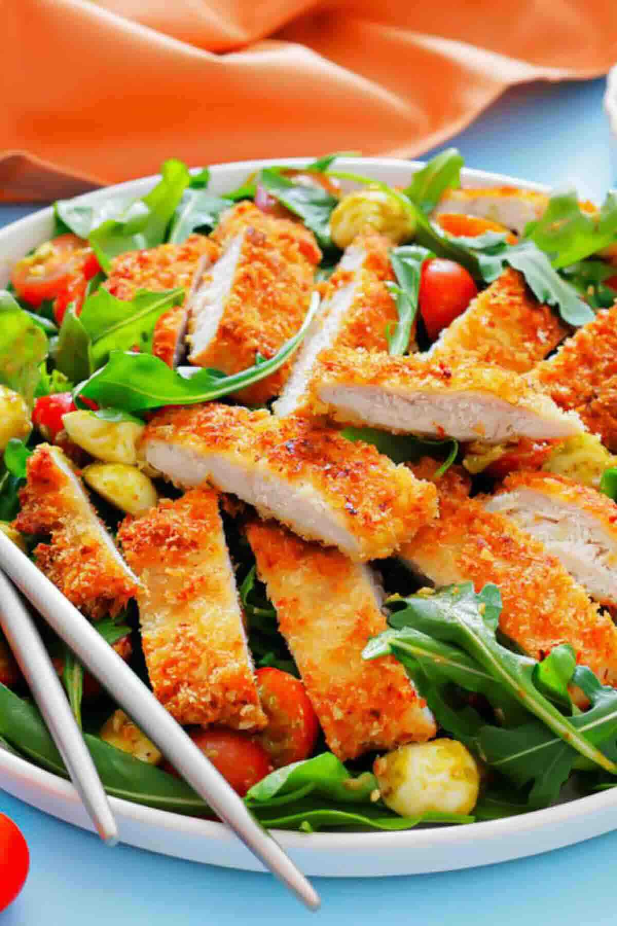 A plate of sliced breaded chicken atop a bed of arugula, cherry tomatoes, and mozzarella balls, with a pair of chopsticks on the side.