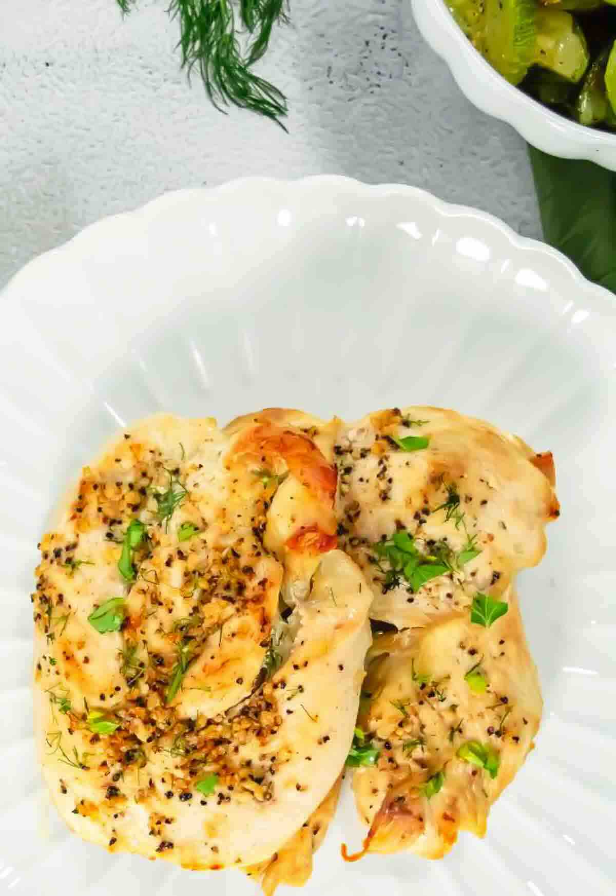 Two pieces of cooked, seasoned chicken breast garnished with herbs are served on a white plate, with a side of vegetables visible in a separate dish nearby.