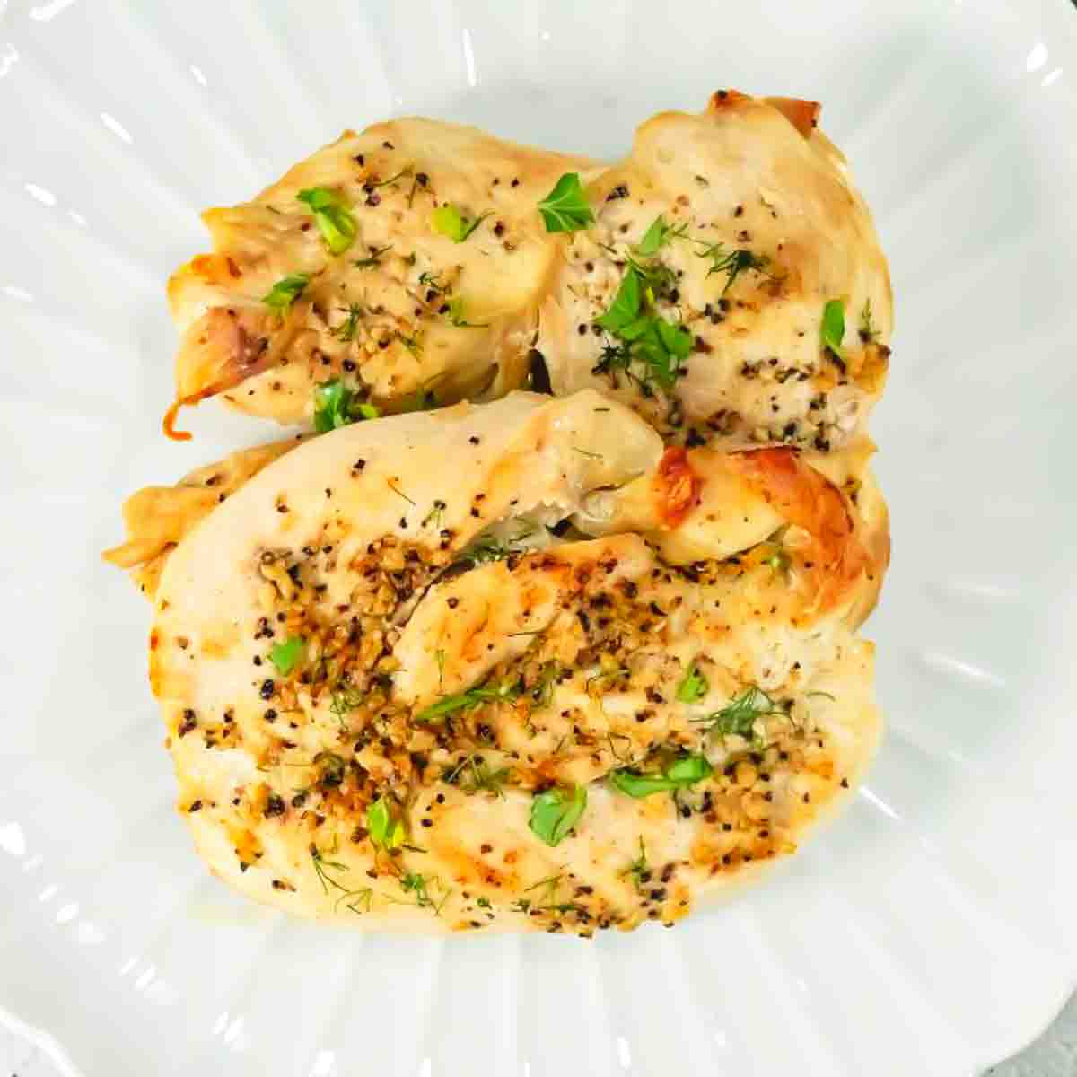 Two baked chicken breasts garnished with chopped herbs and cracked black pepper, served on a white plate.