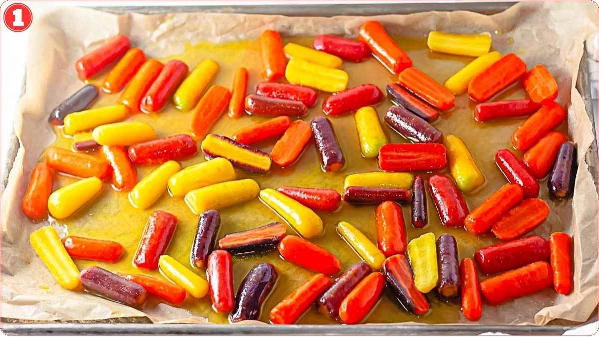 Baking tray with parchment paper holding red, orange, yellow, and purple baby carrots coated in oil or sauce, ready for roasting.