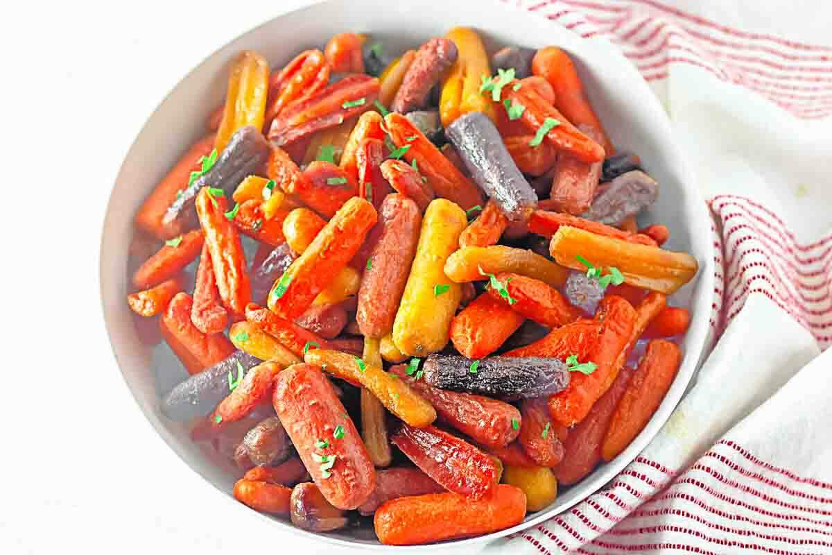A white bowl filled with roasted multicolored baby carrots, garnished with chopped herbs, sits on a surface with a red and white striped cloth nearby.