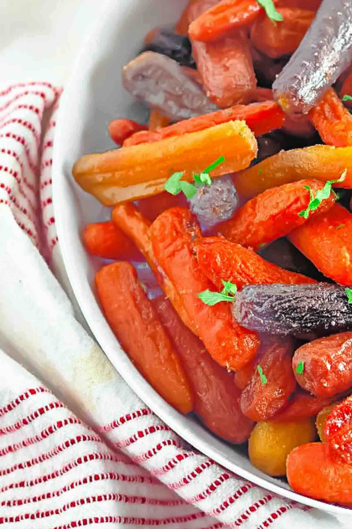 A bowl of cooked multicolored baby carrots garnished with chopped herbs, placed on a white and red striped cloth.