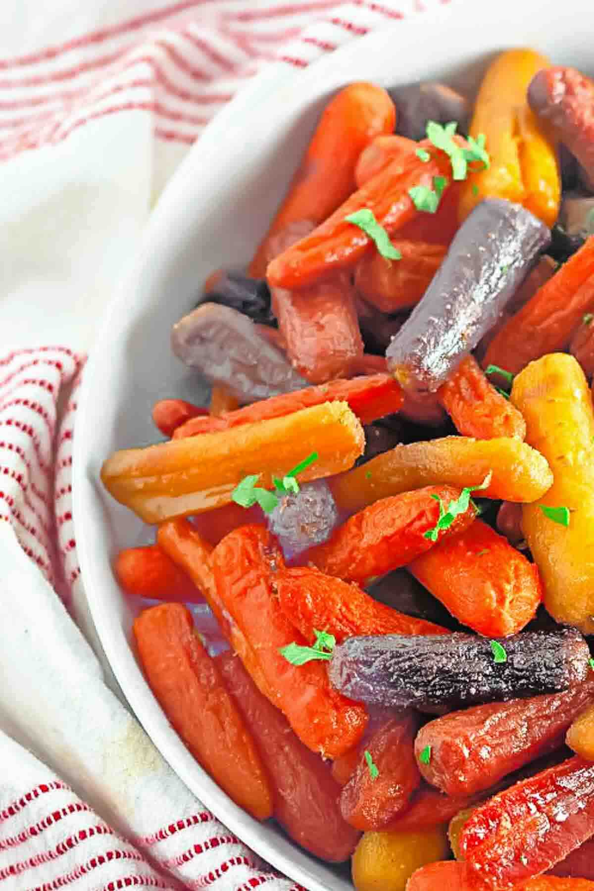Close-up of a bowl filled with roasted multicolored baby carrots, garnished with chopped parsley, on a red-and-white striped cloth.