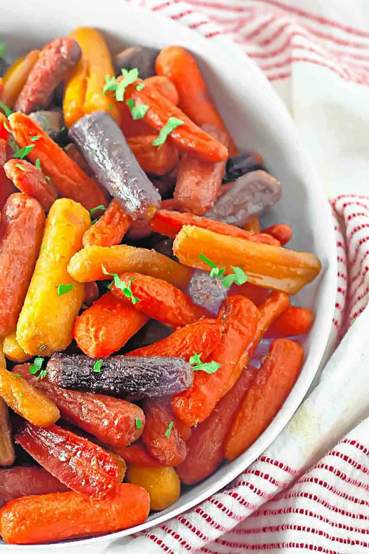 A white bowl filled with steamed multicolored baby carrots, garnished with chopped parsley, sits on a striped red and white cloth.