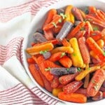 A white bowl filled with assorted colorful baby carrots, garnished with chopped parsley, sits on a red and white striped cloth.