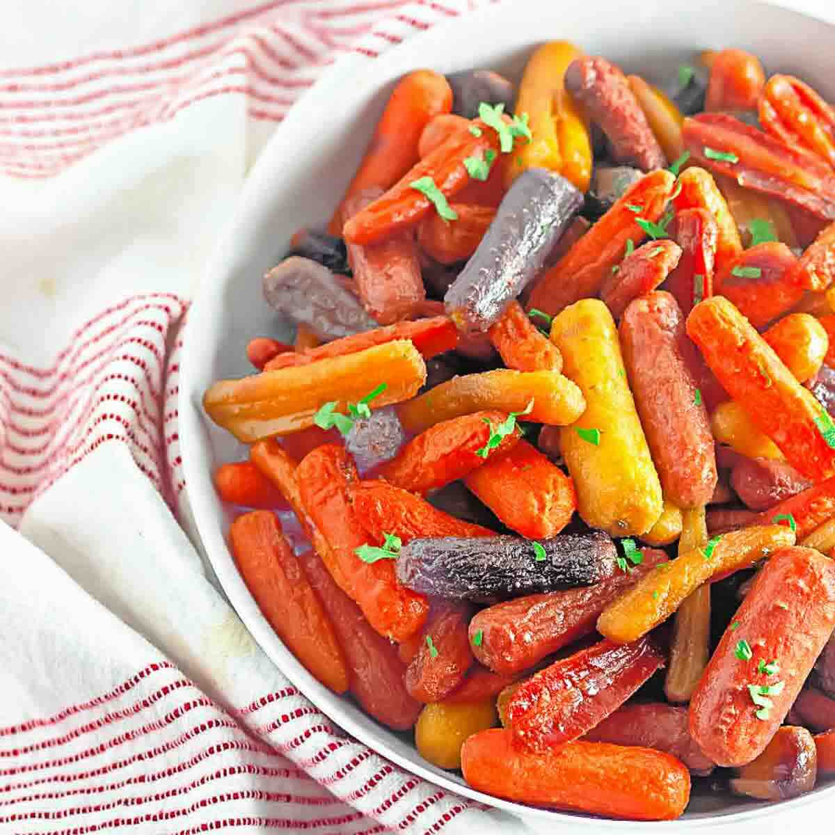 A white bowl filled with assorted colorful baby carrots, garnished with chopped parsley, sits on a red and white striped cloth.