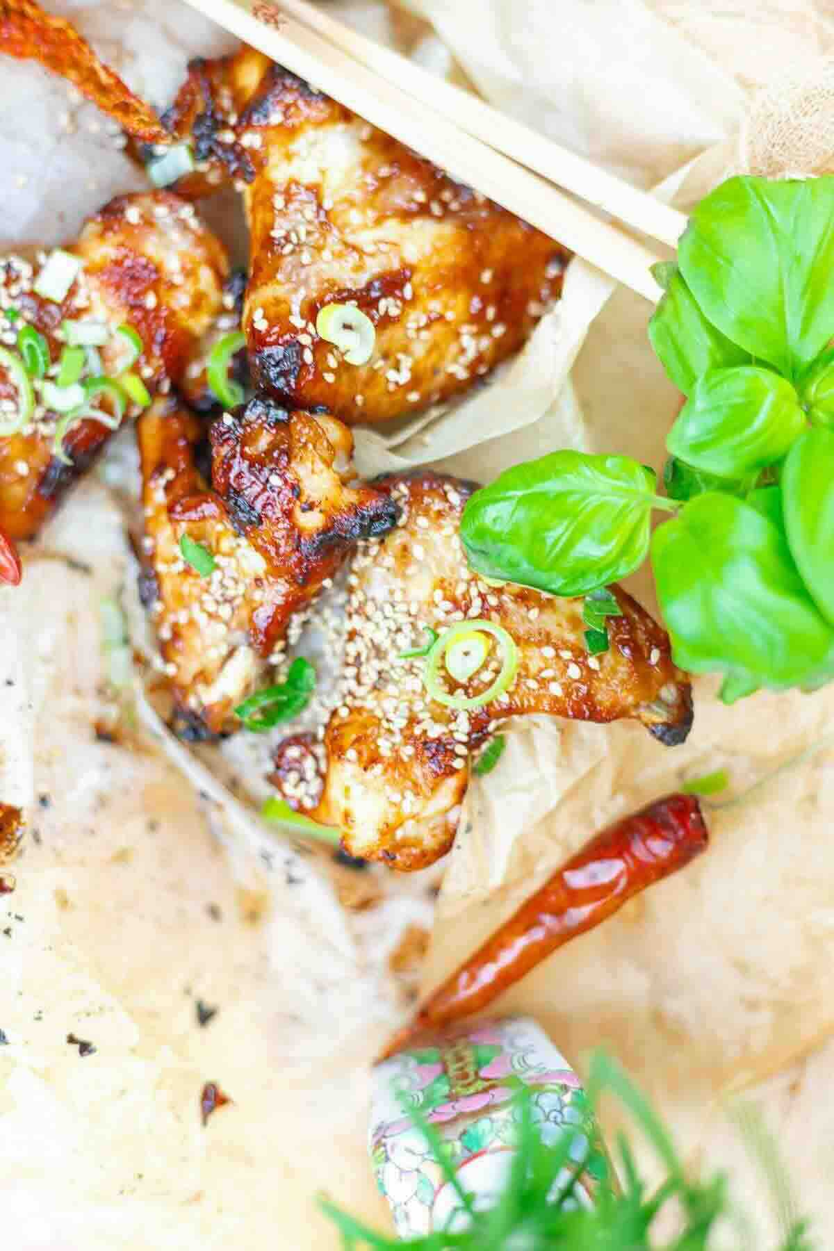 Grilled chicken wings with sesame seeds and green onions, garnished with fresh basil and a red chili, served on parchment with chopsticks.