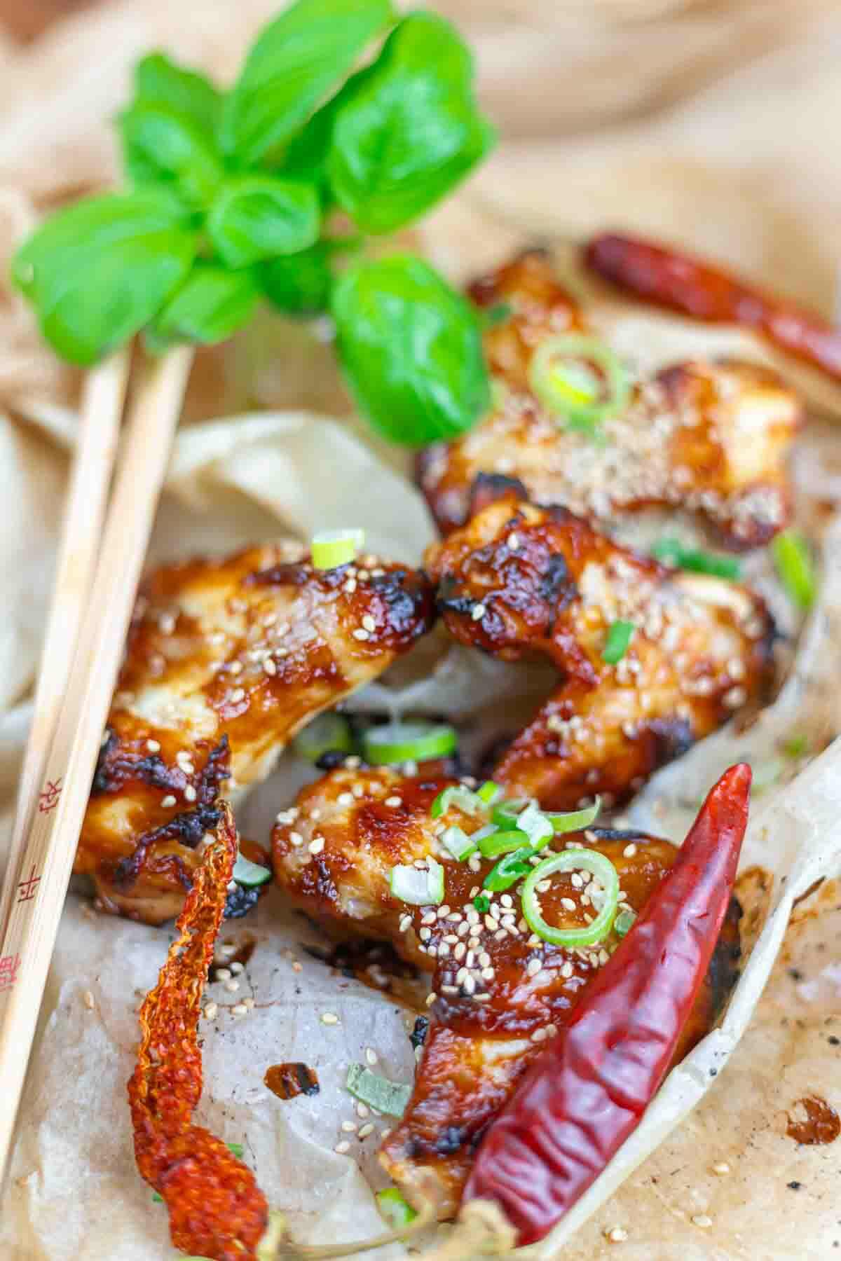 Grilled chicken wings garnished with sesame seeds, green onions, and dried red chili peppers, served with chopsticks and a sprig of fresh herbs.