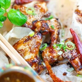 Close-up of glazed chicken wings garnished with sesame seeds, chopped green onions, red chili peppers, and a sprig of fresh basil.