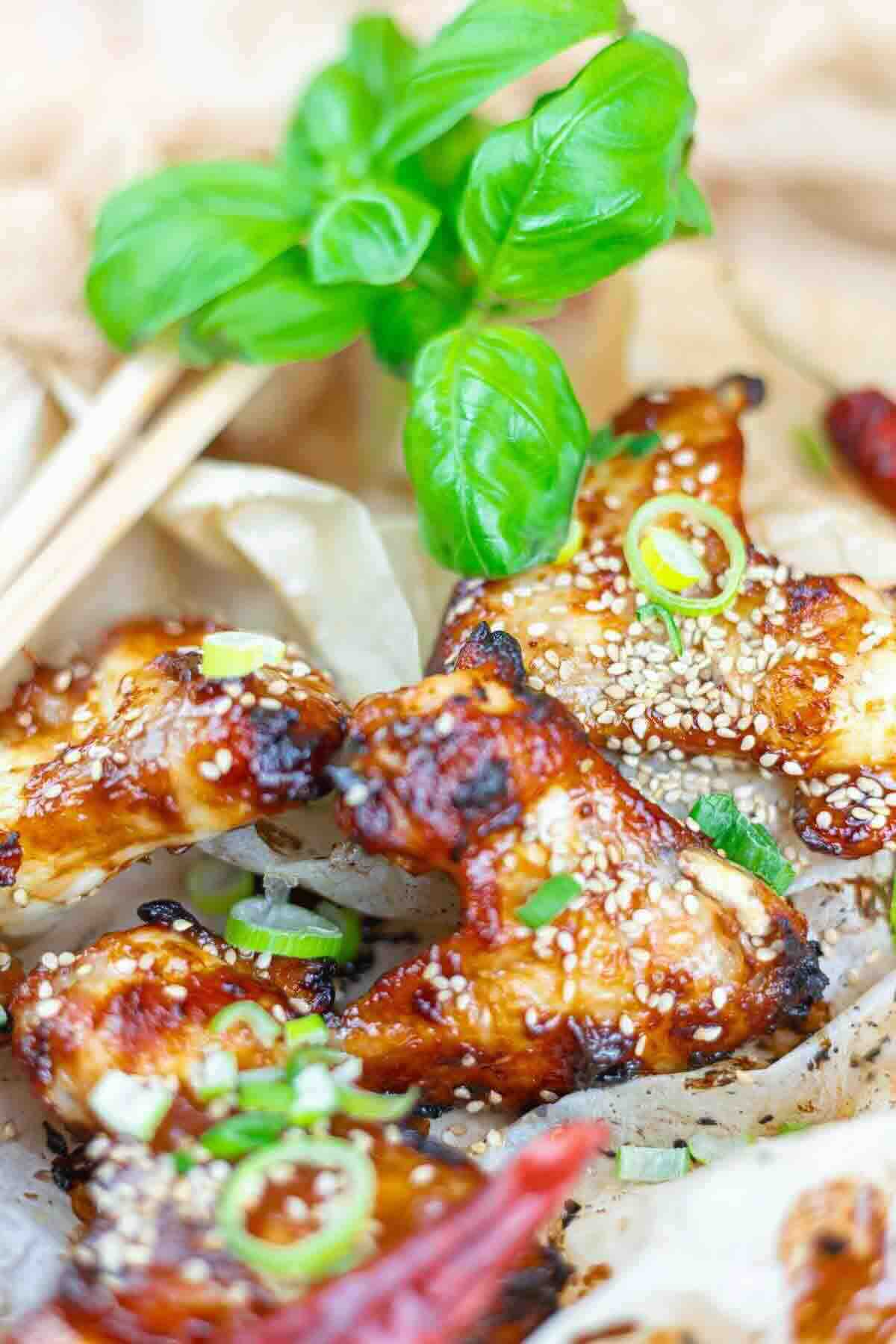Grilled chicken wings garnished with sesame seeds, chopped green onions, and basil leaves, served on parchment paper with chopsticks in the background.