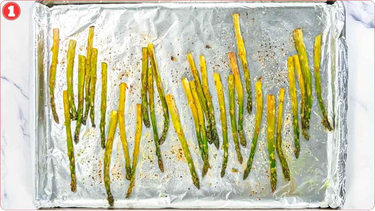 Roasted asparagus spears spread out on a foil-lined baking sheet with light seasoning visible.