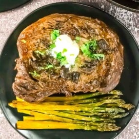 Cooked steak topped with a dollop of butter and chopped herbs, served with roasted asparagus and carrot strips on a black plate.