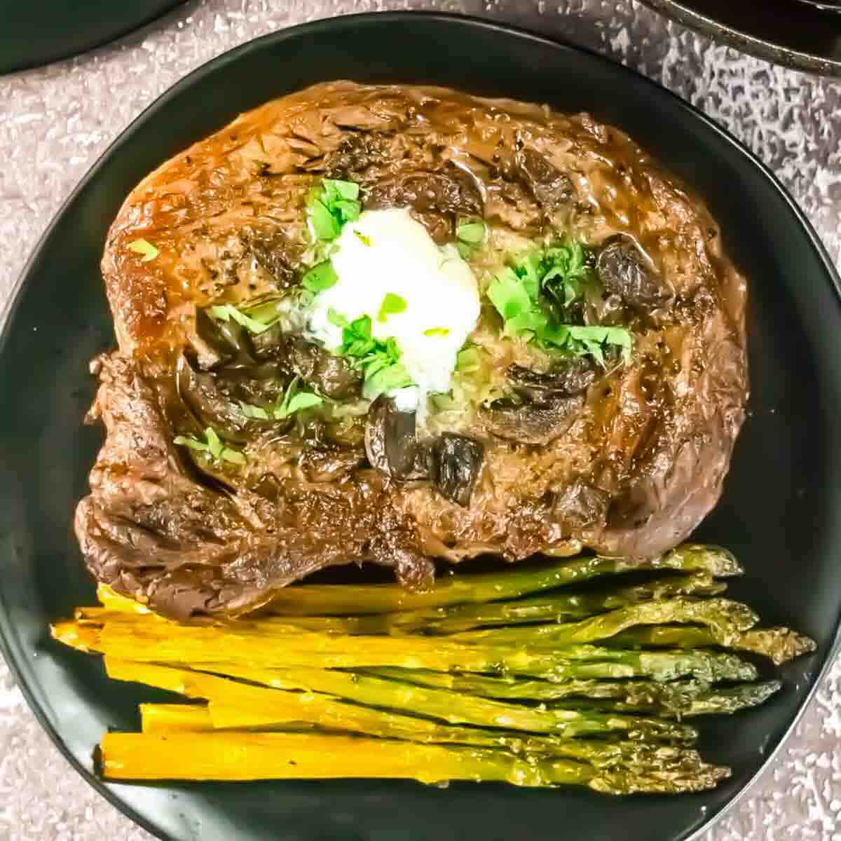 Cooked steak topped with a dollop of butter and chopped herbs, served with roasted asparagus and carrot strips on a black plate.