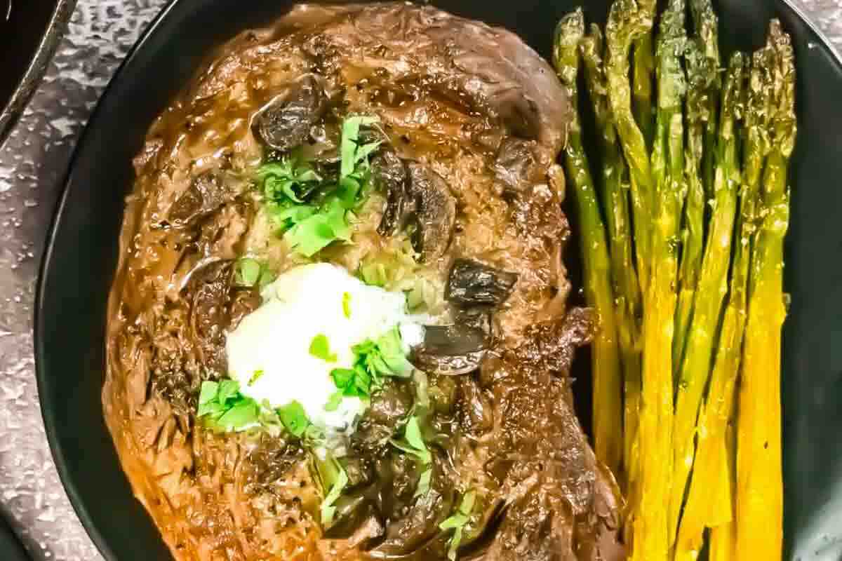 A cooked steak topped with herbs and butter is served on a black plate next to a portion of roasted asparagus.