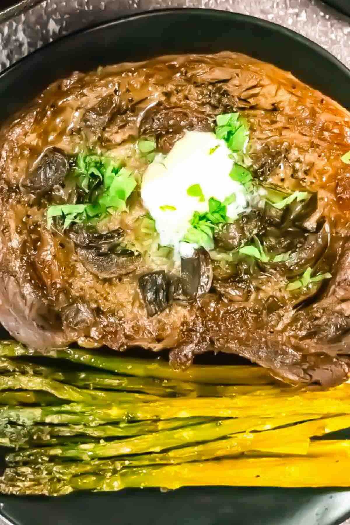 Cooked steak topped with butter and herbs, served with roasted asparagus on a black plate.