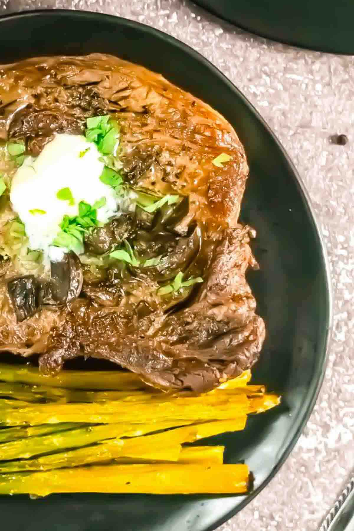 A cooked steak topped with herbs and a dollop of butter, served with yellow roasted asparagus on a black plate.