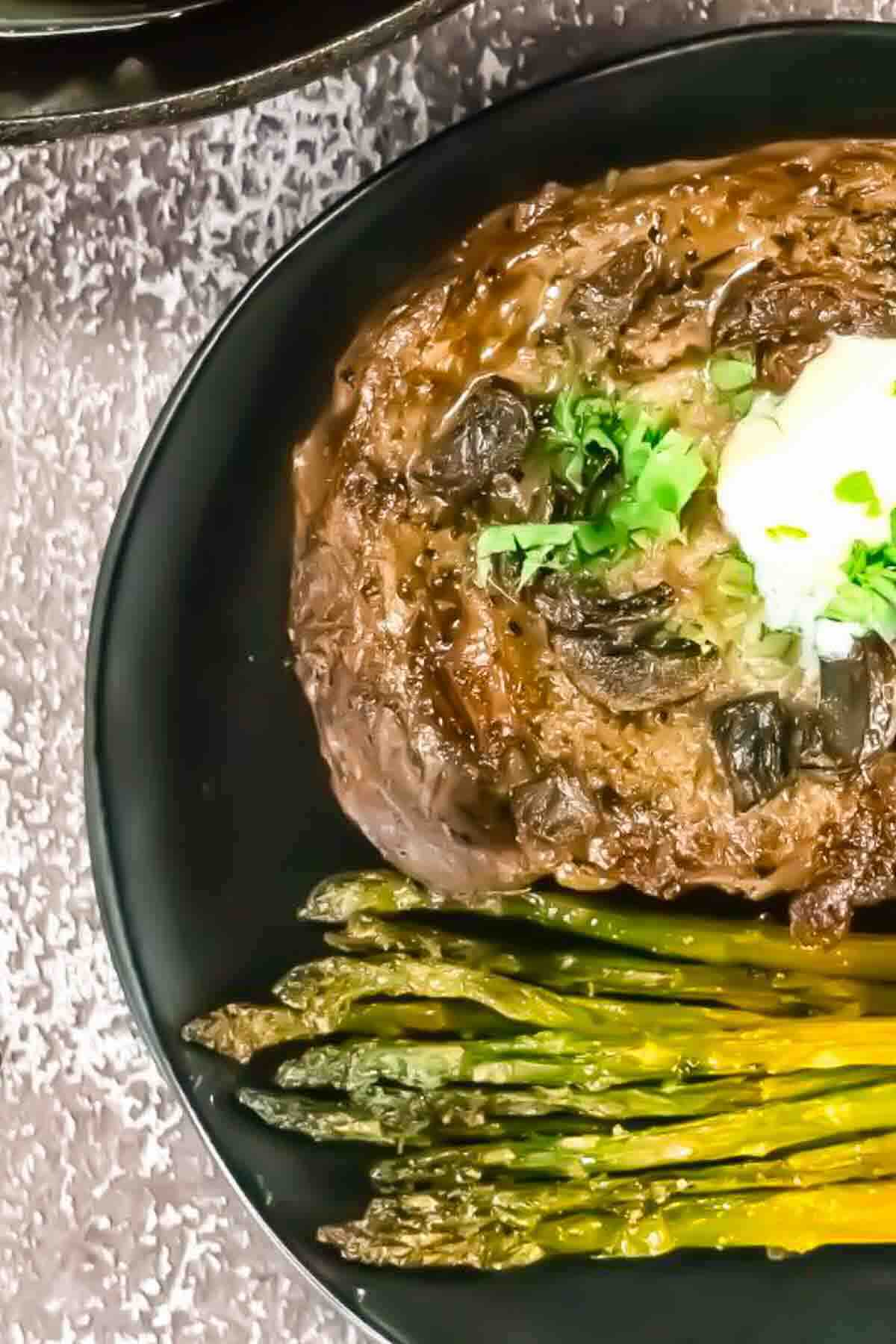 A black plate with a cooked steak topped with herbs and butter, served with roasted asparagus spears on the side.