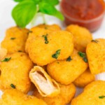 A plate of golden chicken nuggets garnished with herbs, with one nugget cut open to show the inside. A cup of red dipping sauce and basil leaves are in the background.