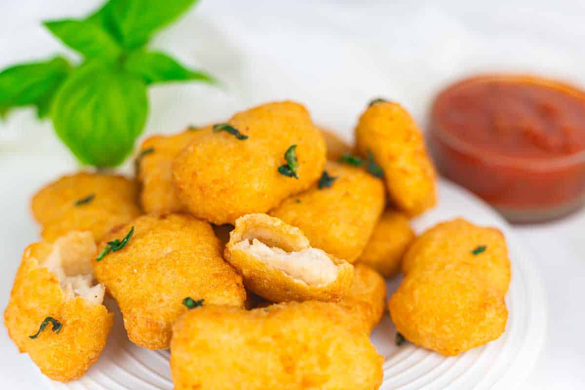 A plate of breaded chicken nuggets garnished with herbs, with a cup of red dipping sauce and a sprig of basil in the background.