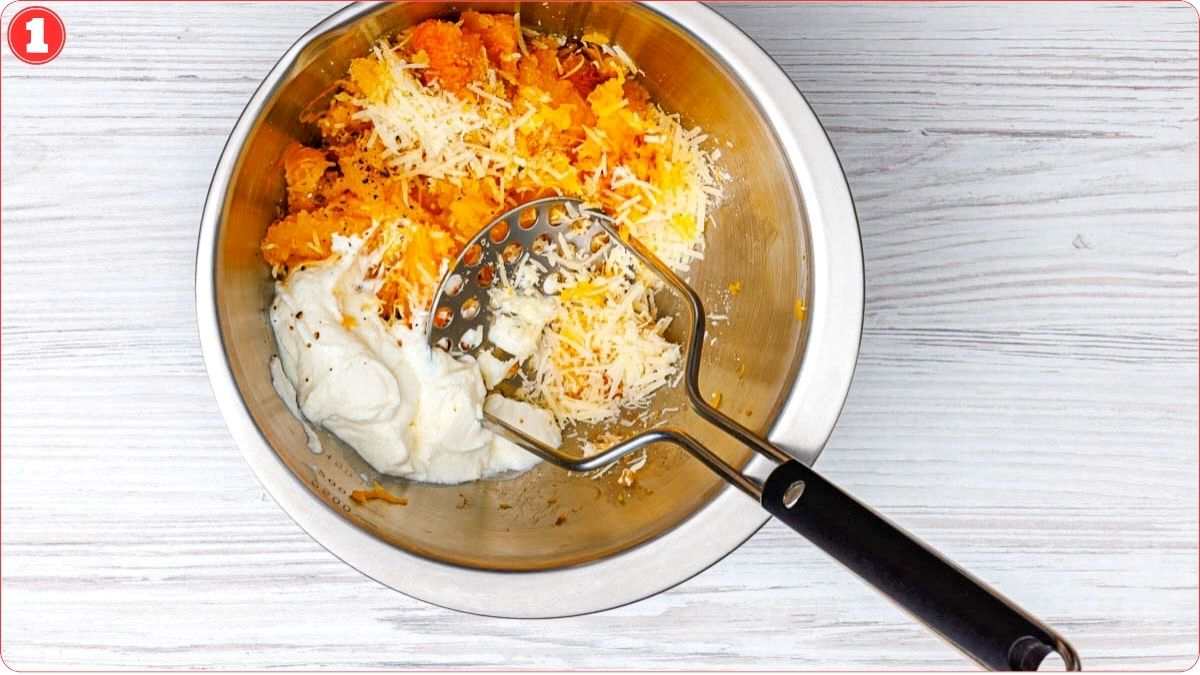 A metal bowl with mashed sweet potatoes, sour cream, shredded cheese, and a potato masher on a white wooden surface.