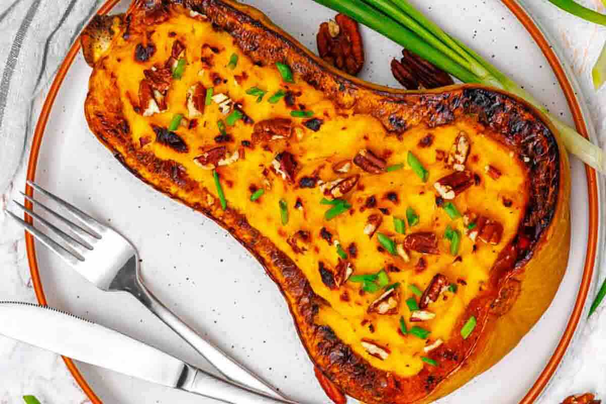 A roasted butternut squash half filled with a creamy, baked cheese mixture, topped with chopped pecans and green herbs, served on a plate with a fork and knife.