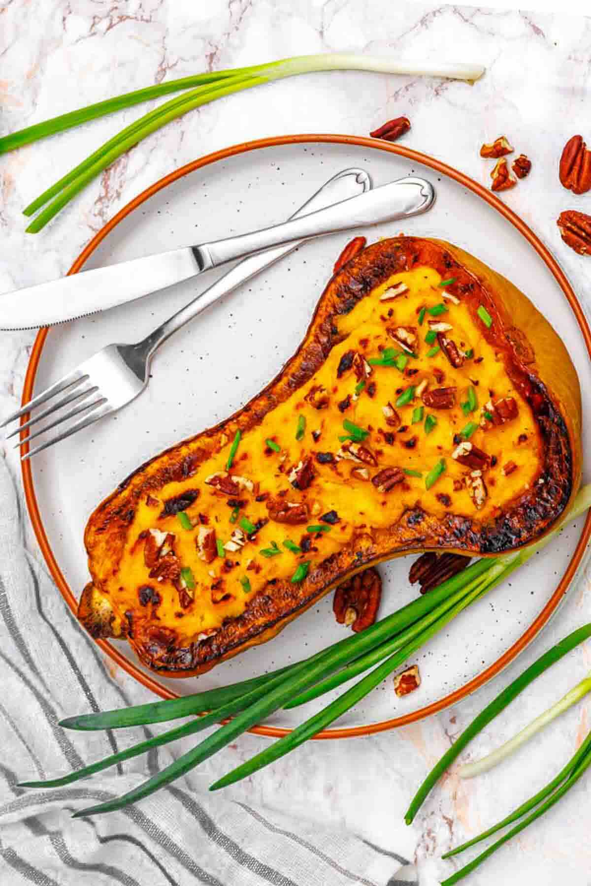 Roasted butternut squash half topped with chopped pecans and chives, served on a plate with a fork, knife, and fresh green onions on the side.